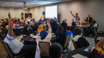 May contain many people in a room with some raising their hands and flags on tables.