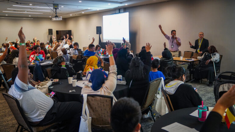 May contain many people in a room with some raising their hands and flags on tables.